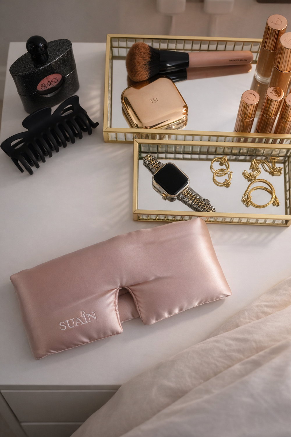Pink Still Suain silk sleep mask with jewellery and beauty products on a bedside table