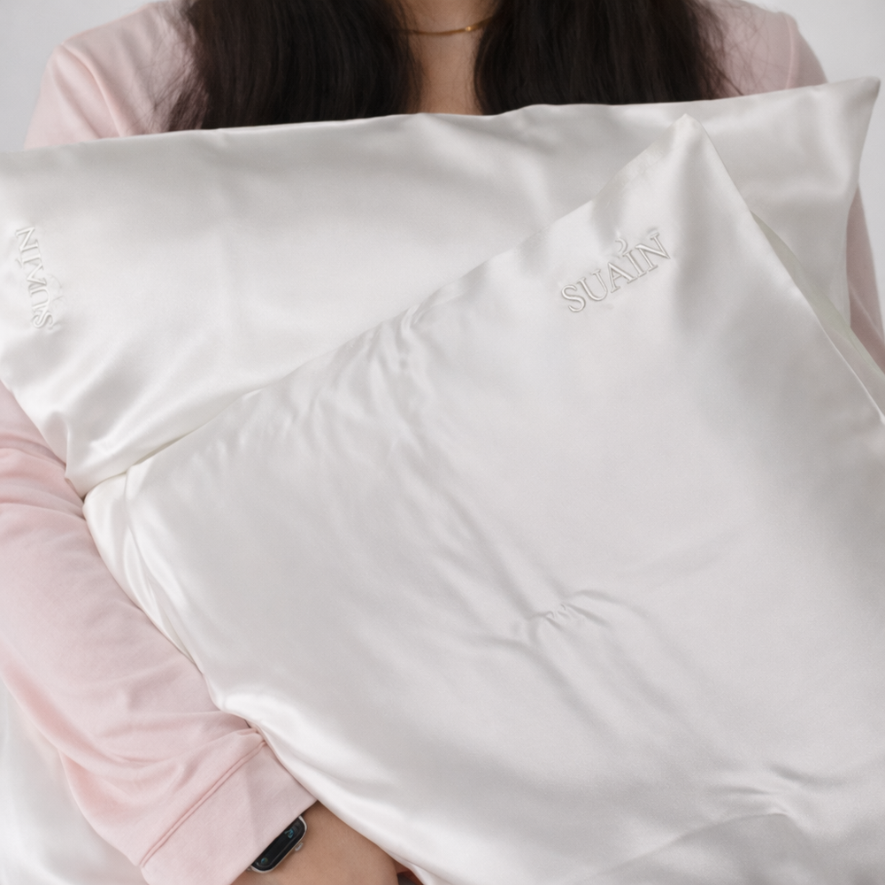 Two Ivory White Still Suain silk pillowcases with the embroidered Suain logo, held in a woman's arms